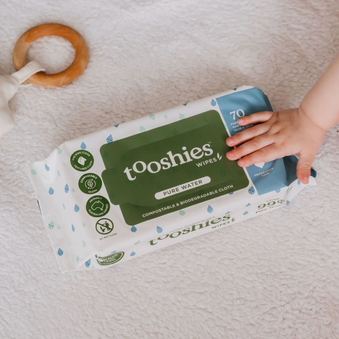 Child's hand holding a package of tooshies wipes on a soft surface with a wooden toy nearby.